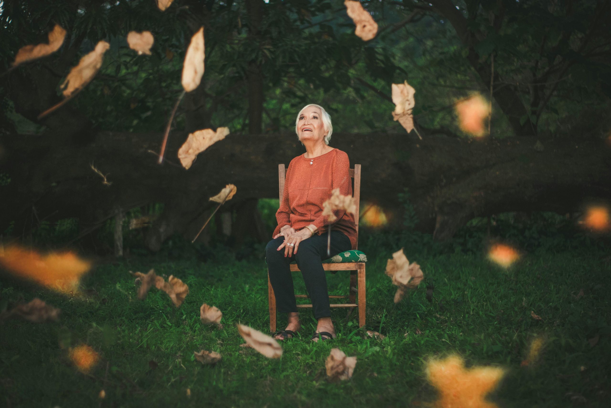 An elderly woman sits outdoors enjoying the falling autumn leaves, capturing a serene moment.